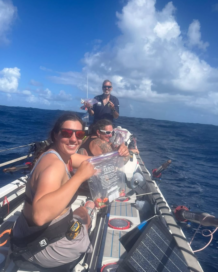 Team of rowers in the middle of the Atlantic smiling and opening rations to eat